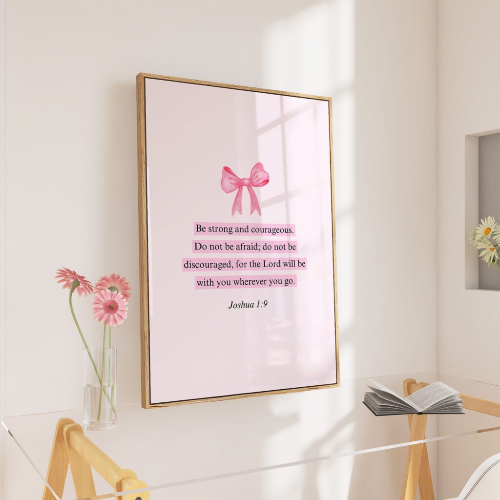 Framed quote with a pink bow, next to a vase with pink flowers on a clear desk. An open book is on the desk.